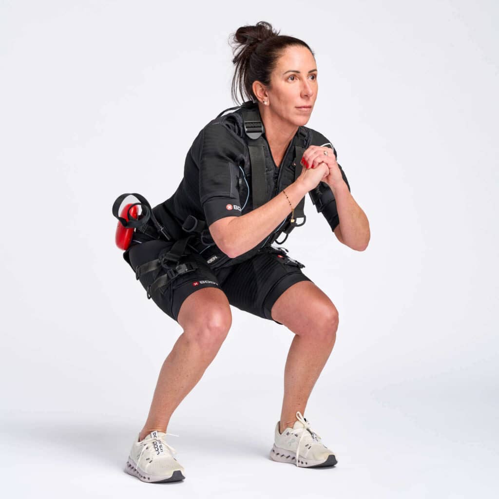 SOMA Fit Club A woman wearing a black EMS training suit performs a squat exercise against a plain white background, showcasing the efficiency of SOMA Fit Club’s 20 Minute workout. EMS Training