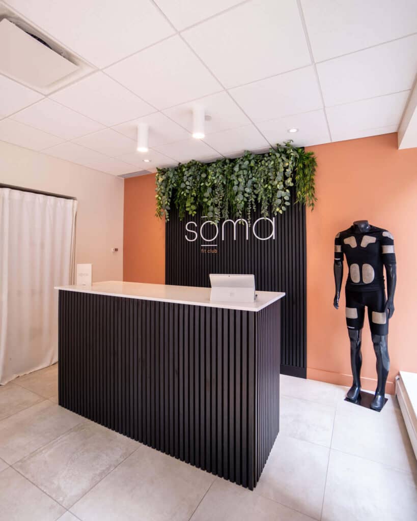 SOMA Fit Club Reception area with a black front desk, a laptop, green plant wall featuring the “soma” sign, an orange accent wall, and a mannequin in a black suit with white patches promoting our 20 Minute EMS workout at SOMA Fit Club. EMS Training