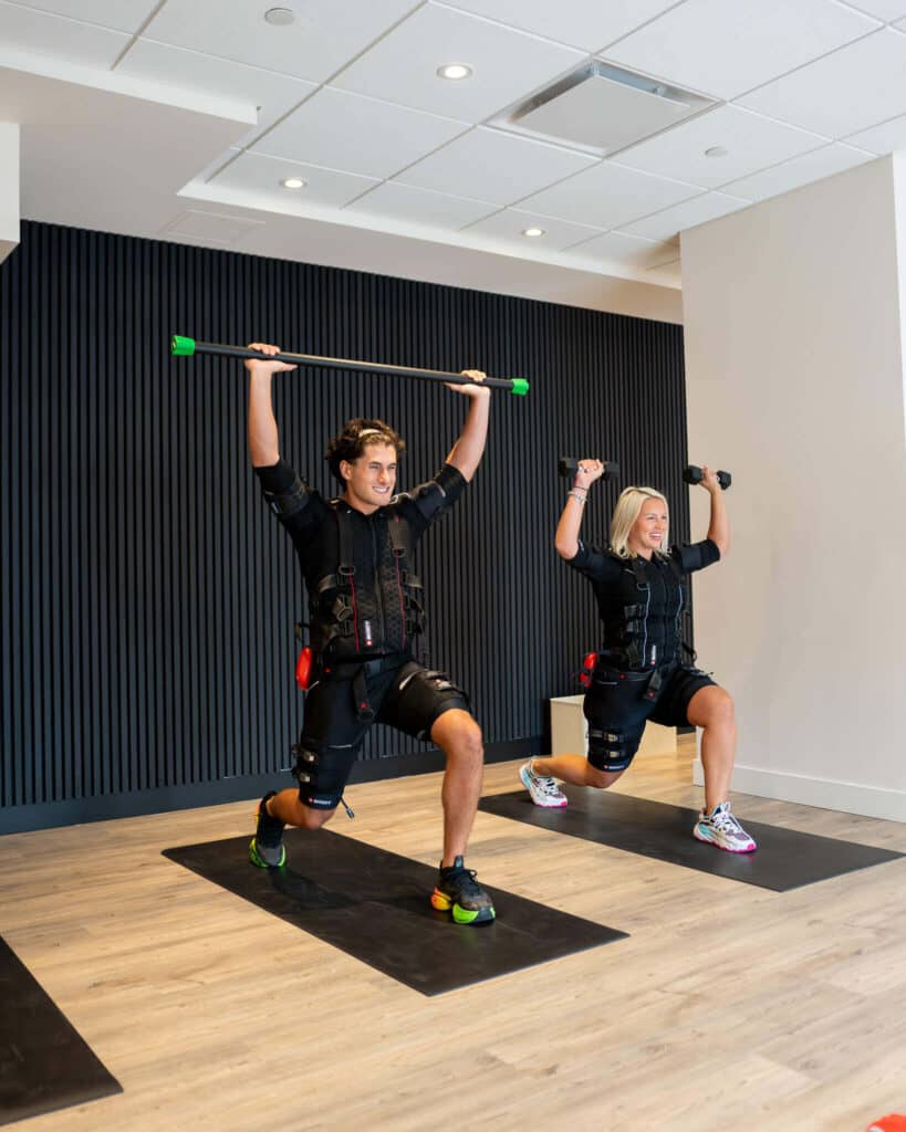 SOMA Fit Club Two people at SOMA Fit Club do lunges on mats indoors in an EMS workout; one lifts a barbell overhead, the other holds dumbbells—all as part of an intense 20 minute workout. EMS Training