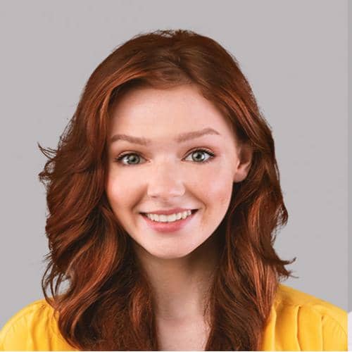 SOMA Fit Club A young woman with red hair and green eyes smiles at the camera. She is wearing a yellow top, ready for her 20 Minute workout with EMS Training, and is photographed against a plain light gray background. EMS Training