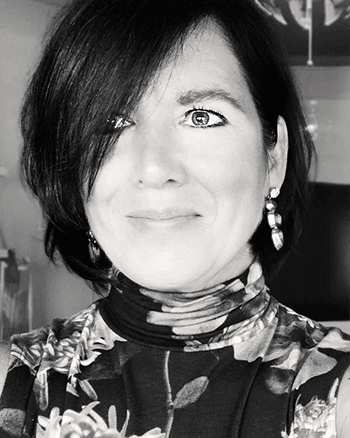 SOMA Fit Club A woman with short dark hair, wearing a floral turtleneck top and earrings, looks at the camera and smiles slightly. The black-and-white photo captures her at SOMA Fit Club after an energizing EMS workout. EMS Training