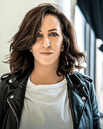 SOMA Fit Club Woman with shoulder-length brown hair in a white shirt and black leather jacket, standing indoors near a window with natural light, ready for her 20 Minute workout at SOMA Fit Club. EMS Training