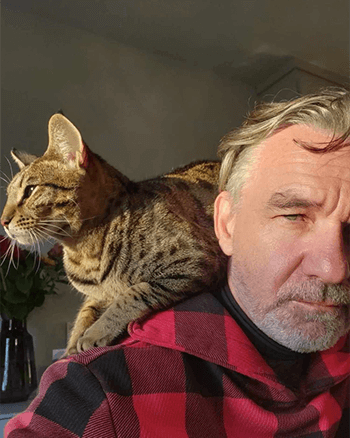 SOMA Fit Club A man in a red plaid shirt stands indoors with a tabby cat perched on his shoulder, both looking in the same direction, as if ready to head out for an EMS workout at SOMA Fit Club. EMS Training