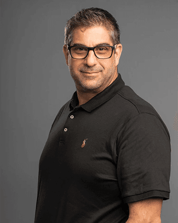 SOMA Fit Club A man with short dark hair and glasses wearing a black polo shirt stands against a plain gray background, representing EMS Training and the benefits of a 20 Minute workout from EMS Canada as he looks at the camera. EMS Training