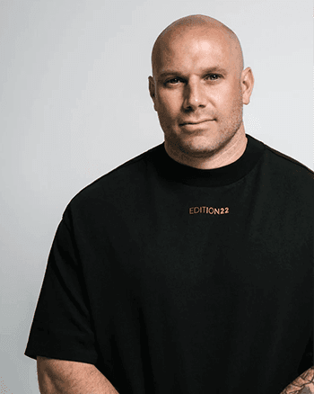 SOMA Fit Club A bald man wearing a black t-shirt with "EDITION22" stands against a plain light background, looking at the camera with a neutral expression—perfectly capturing the focused energy of an EMS Training or 20 Minute workout session. EMS Training