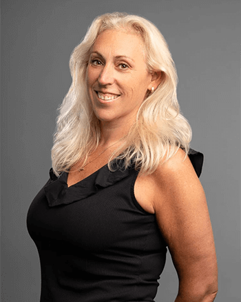 SOMA Fit Club A woman with long, wavy blonde hair is smiling and standing against a plain gray background. She is wearing a sleeveless black top, embodying the energy of a 20 Minute workout inspired by EMS Training from EMS Canada. EMS Training